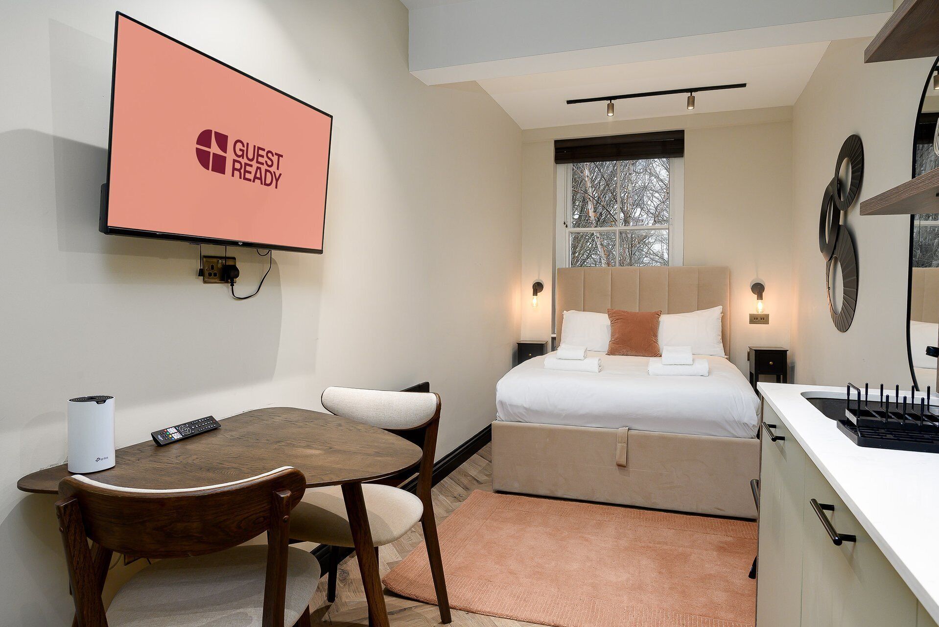 Een hotelkamer met een bed, nachtkastje, bureau met stoel, een aan de muur bevestigde televisie en een raam met uitzicht op bomen.