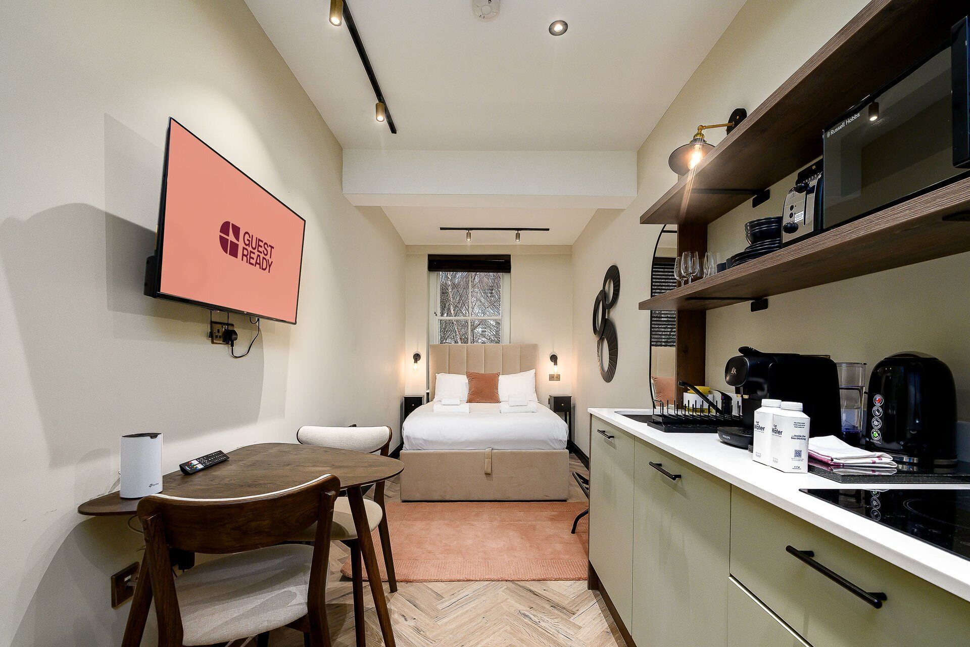 Een compacte hotelkamer met een bed, een klein eettafeltje, een kitchenette en een televisie waarop 'GUEST READY' staat.