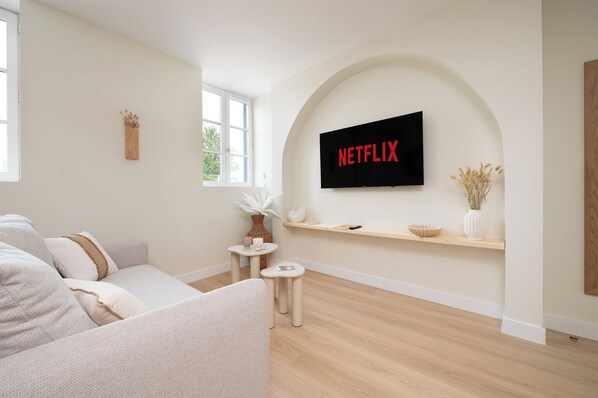 A modern living room with a flat-screen TV mounted on a wall, a sofa, a small side table, and a window with a view of greenery. - Free parking • Downtown on foot • Air Conditioning (Albi)
