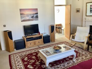 Ein Wohnzimmer mit Fernseher, Lautsprechern, einem Couchtisch, einem roten Teppich mit Muster und einem weißen Sessel.
