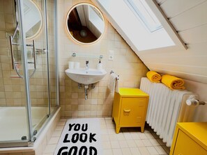 Ein modernes Badezimmer mit Duschkabine aus Glas, einem Waschbecken mit zwei Wasserhähnen, einem Spiegel, einem gelben Schrank und einem Heizkörper.