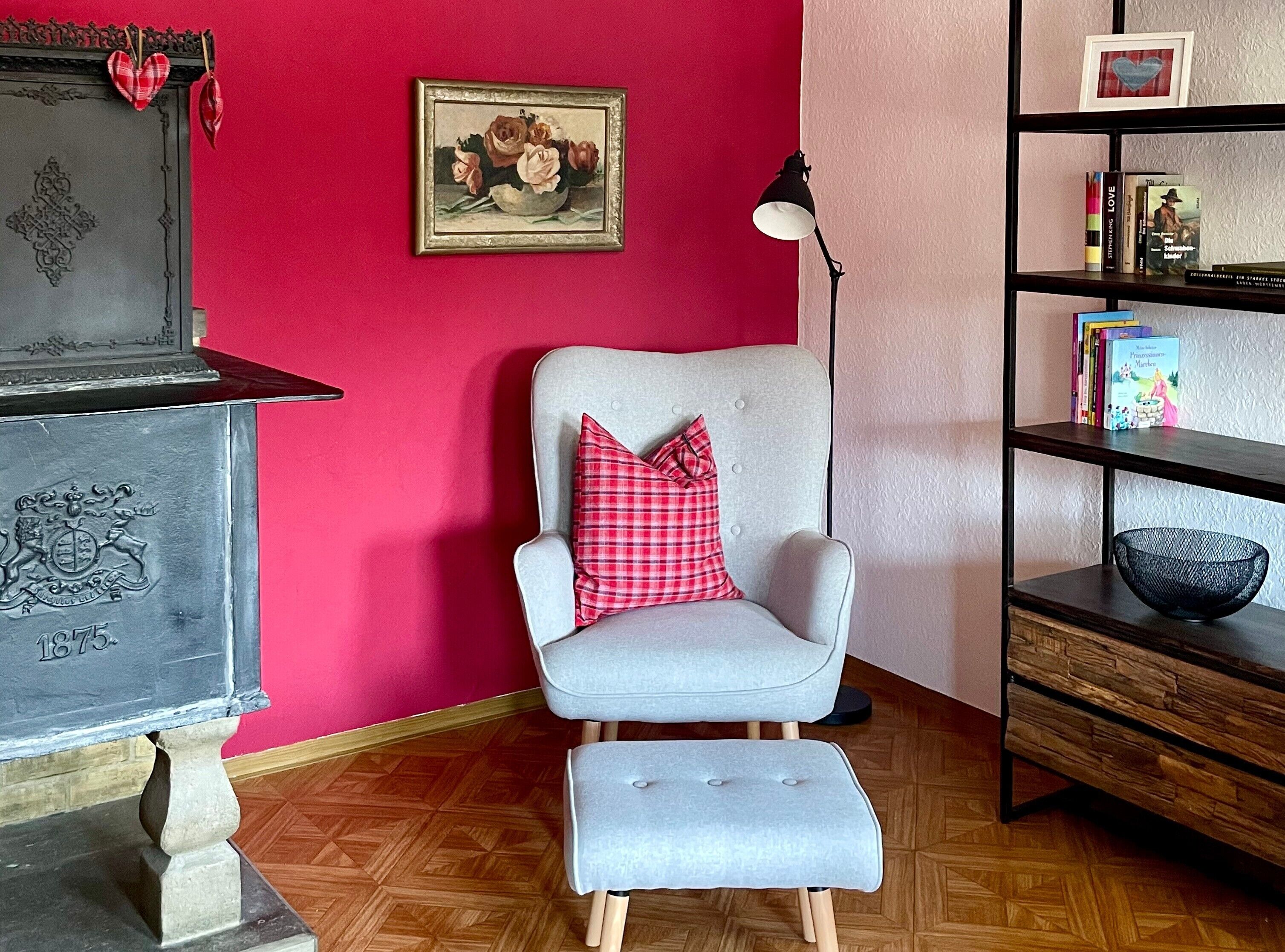A room with a red wall, a grey armchair with a red pillow, a bookshelf, and a decorative plate with a crest.