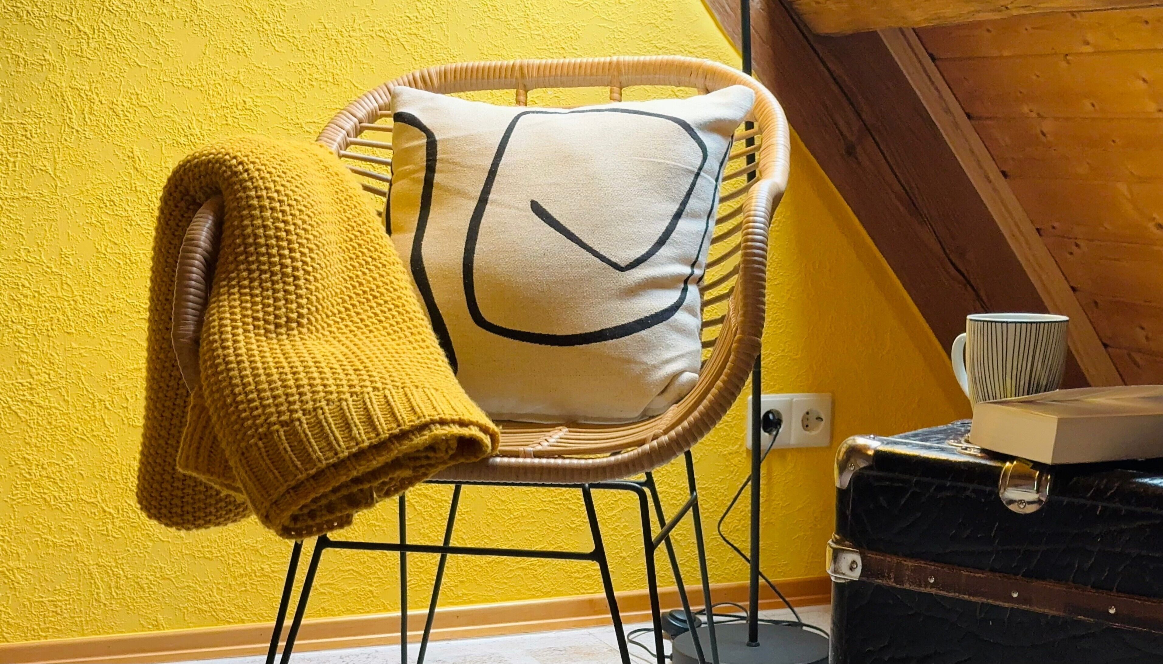 Ein Korbstuhl mit Kissen, eine gelb gestrickte Decke und ein Buch auf einem Tisch vor einer gelben Wand.