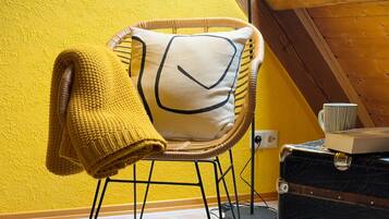 A wicker chair with a cushion, a yellow knitted throw, and a book on a table against a yellow wall.