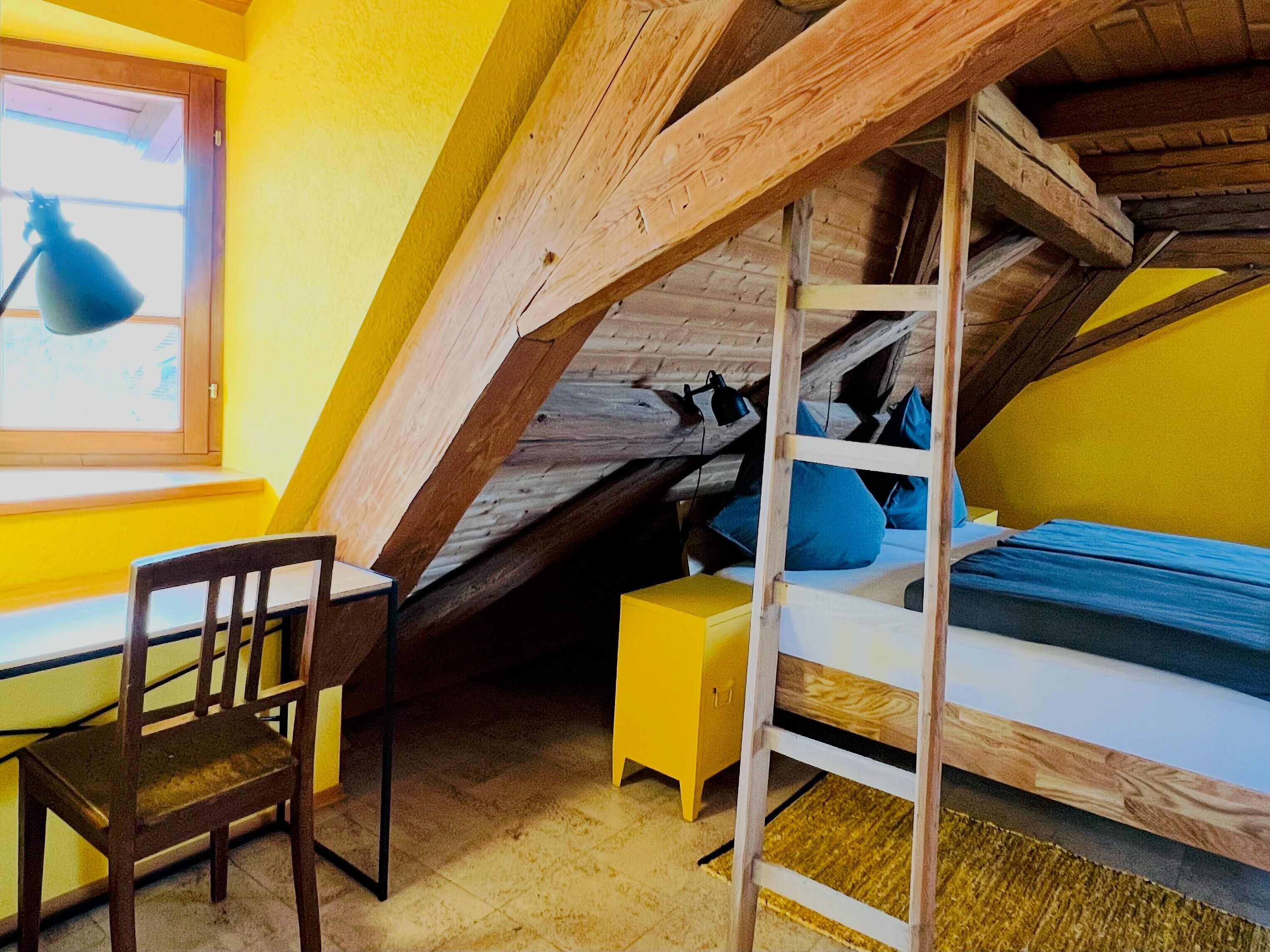 A room with a sloped ceiling, a bed with blue bedding, a wooden chair, and a small yellow bedside table.