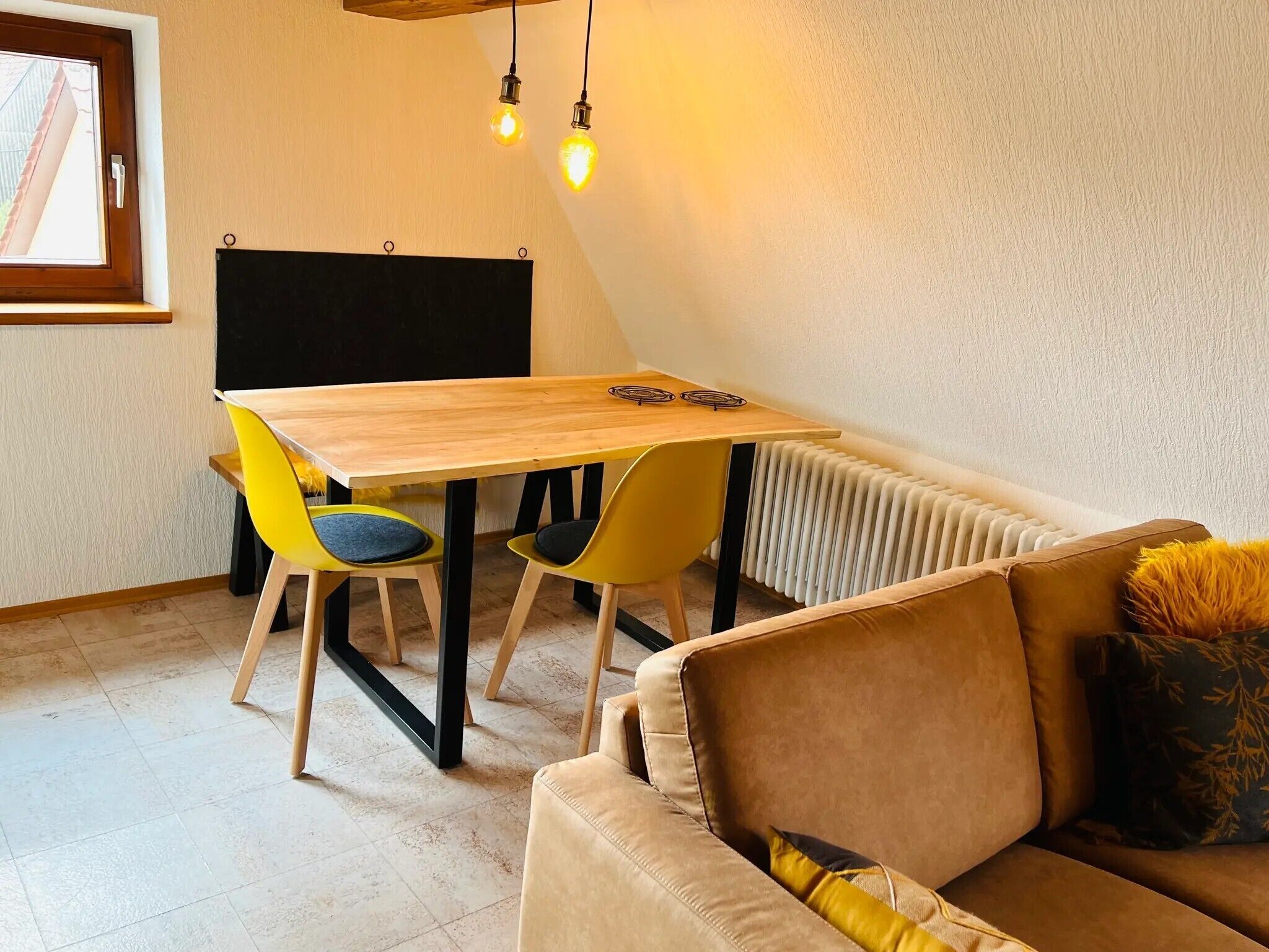 A small, cozy room with a wooden table, two yellow chairs, a brown sofa, and a radiator.