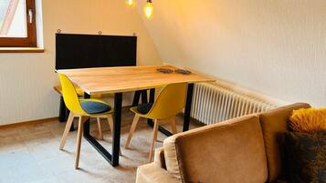 A small, cozy room with a wooden table, two yellow chairs, a brown sofa, and a radiator.