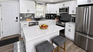 Une cuisine moderne avec des meubles blancs, un réfrigérateur en acier inoxydable, un four à micro-ondes et un îlot central agrémenté d’un panier de fruits.