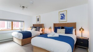 Quarto de hotel com duas camas, uma janela, um radiador e uma mesa de cabeceira com abajur.