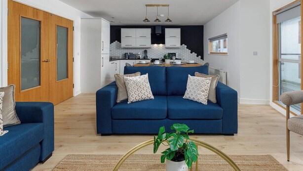 Uma sala de estar moderna com um sofá azul, uma mesa de centro com uma planta e uma cozinha ao fundo.