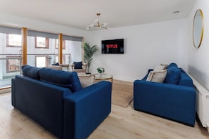 Ein modernes Wohnzimmer mit zwei blauen Sofas, einem an der Wand montierten Fernseher und einem großen Fenster mit Blick ins Freie.