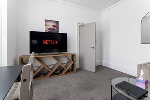 Ein Zimmer mit Fernseher auf einem Holzboden, einer Tür, einem Glastisch mit einem Buch und einer kleinen Pflanze.