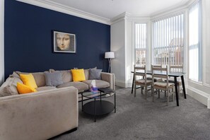 A living room with a dark blue wall, a beige sofa with yellow cushions, a round coffee table, and a dining area with a table and chairs. - Flatzy- Sleeps 16 Near Football Ground + Ping Pong (Liverpool)