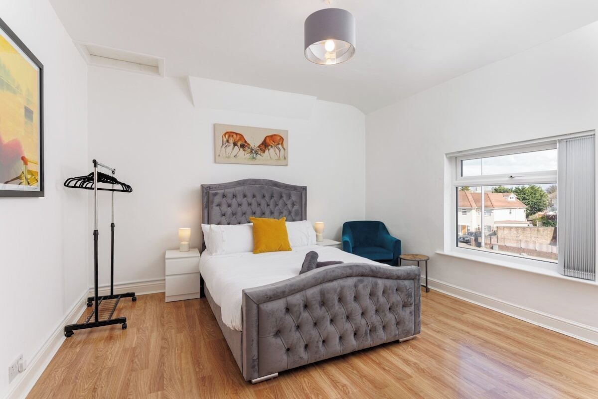 Een slaapkamer met een grijs, gestoffeerd bed, een nachtkastje met een lamp, een stoel, een vloerlamp en een raam met uitzicht.