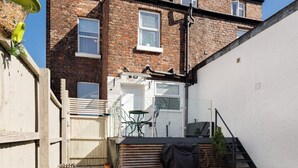 Ein Wohngebäude mit Backsteinfassade, einer weißen Seitenwand, einem Holzzaun, einem kleinen Garten mit Rasen und einer Treppe, die zu einer Terrasse führt.