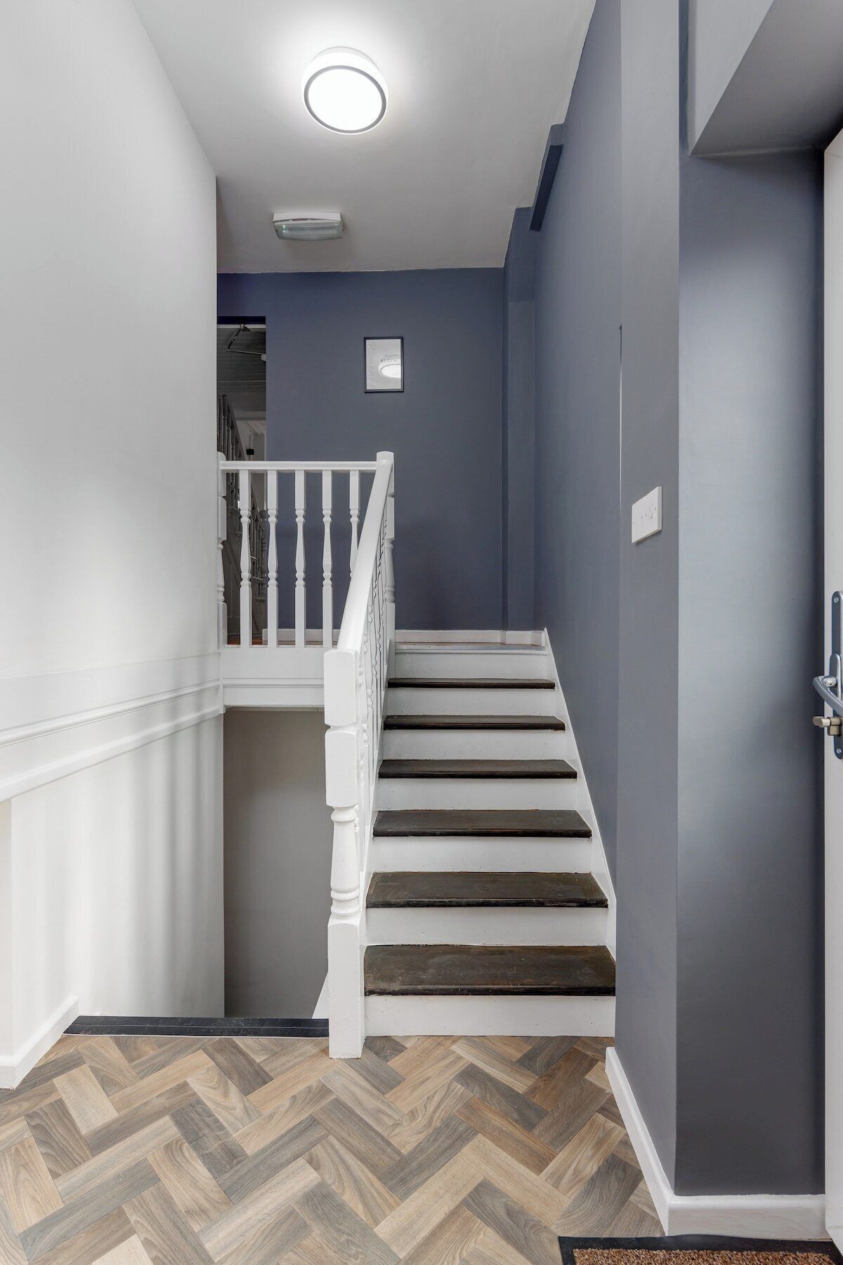 Una escalera con balaustres blancos y peldaños oscuros, que conduce a un rellano con un cuadro enmarcado.
