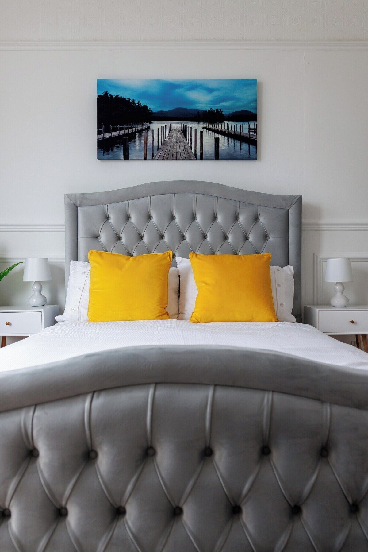 Un dormitorio con cabecera capitonada, cama con ropa blanca y dos almohadas amarillas, y un cuadro de un muelle en la pared.
