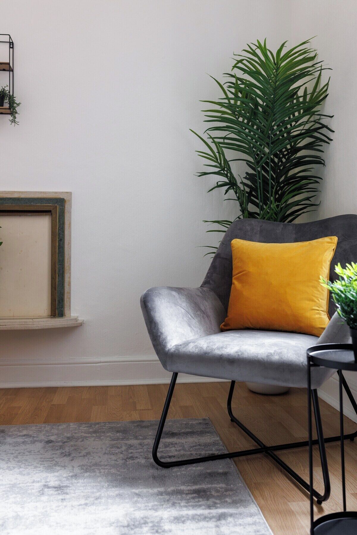 Een moderne woonkamer met een grijze fauteuil, een geel kussen, een grote groene plant en een minimalistische plank.