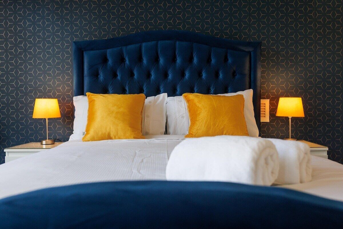 A bed with a tufted headboard, white bedding, and two yellow pillows. There are two bedside lamps on either side of the bed.