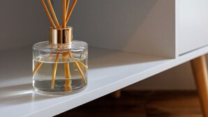 En genomskinlig glasflaska med träpinnar i, placerad på en vit hylla.
