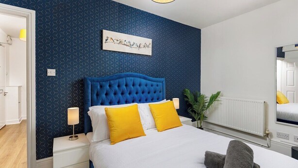 Ein Schlafzimmer mit blauer Wand in geometrischem Muster, einem gepolsterten Kopfteil, weißer Bettwäsche und zwei Nachttischlampen.