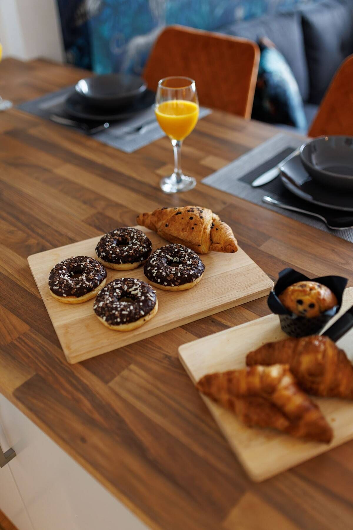 Une table de salle à manger en bois, mise pour le petit-déjeuner, avec des croissants, des beignets enrobés de chocolat et un verre de jus d’orange.