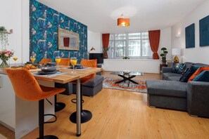 A modern living room with a dining area, featuring a wooden table, orange chairs, a grey sofa, and a patterned wall. - Flatzy - Spacious City Centre Retreat  Sleeps 6 (Merseyside)