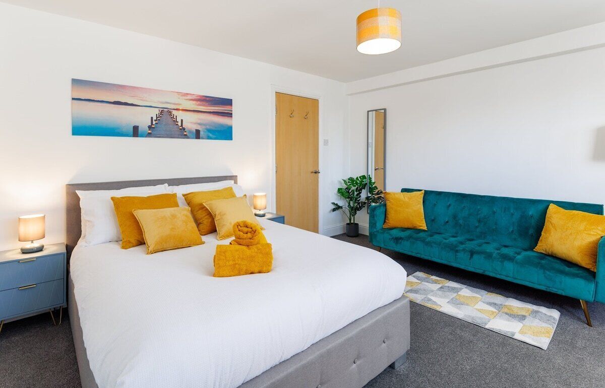 A bedroom with a bed, a nightstand, a teal sofa, a wall-mounted mirror, and a painting.
