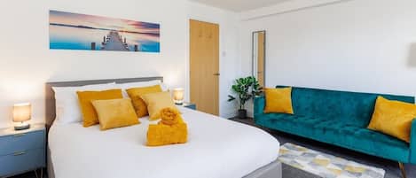 Un dormitorio con cama, mesita de noche, sofá color turquesa, espejo en la pared y un cuadro.