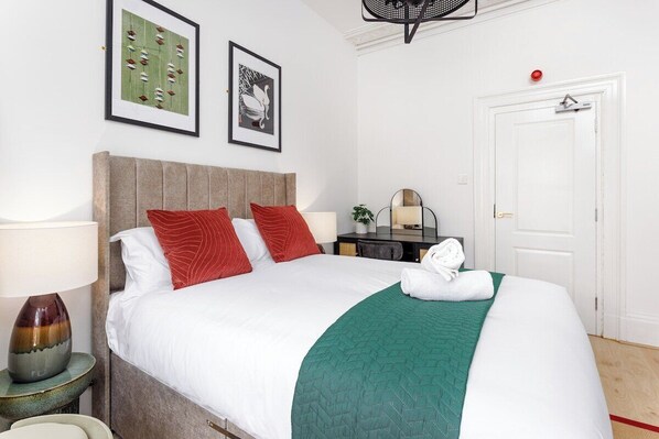 A neatly made bed with a green blanket, a red pillow, and a white pillow. There is a lamp on a bedside table, a framed picture on the wall, and a door with a red handle. - Flatzy–City Centre Mansion 2 | Sleeps 22 | 4 Baths (Liverpool)