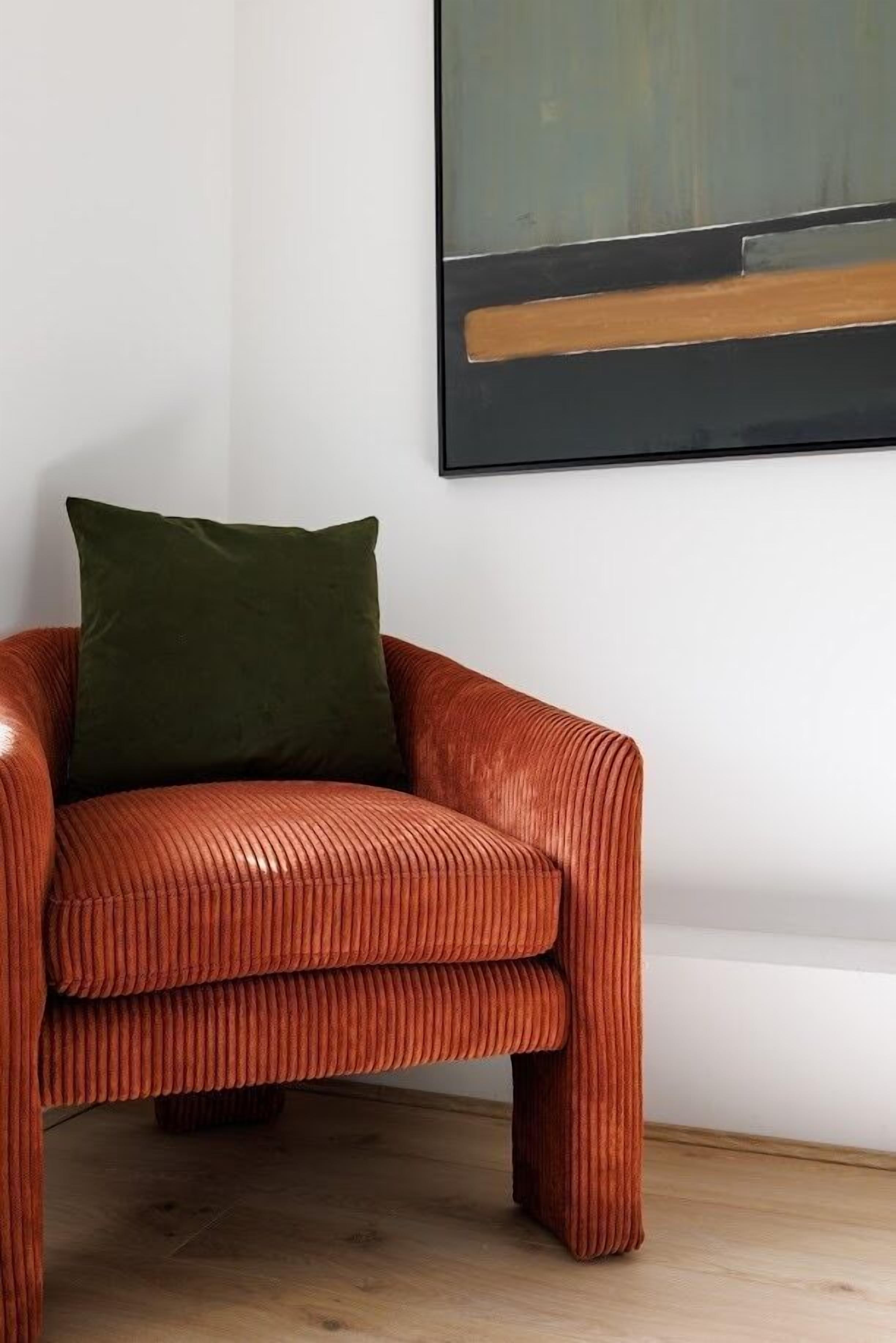 Um sofá de veludo vermelho com uma almofada verde, encostado a uma parede branca, com um quadro pendurado acima.