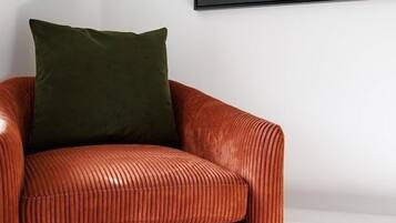 Um sofá de veludo vermelho com uma almofada verde, encostado a uma parede branca, com um quadro pendurado acima.
