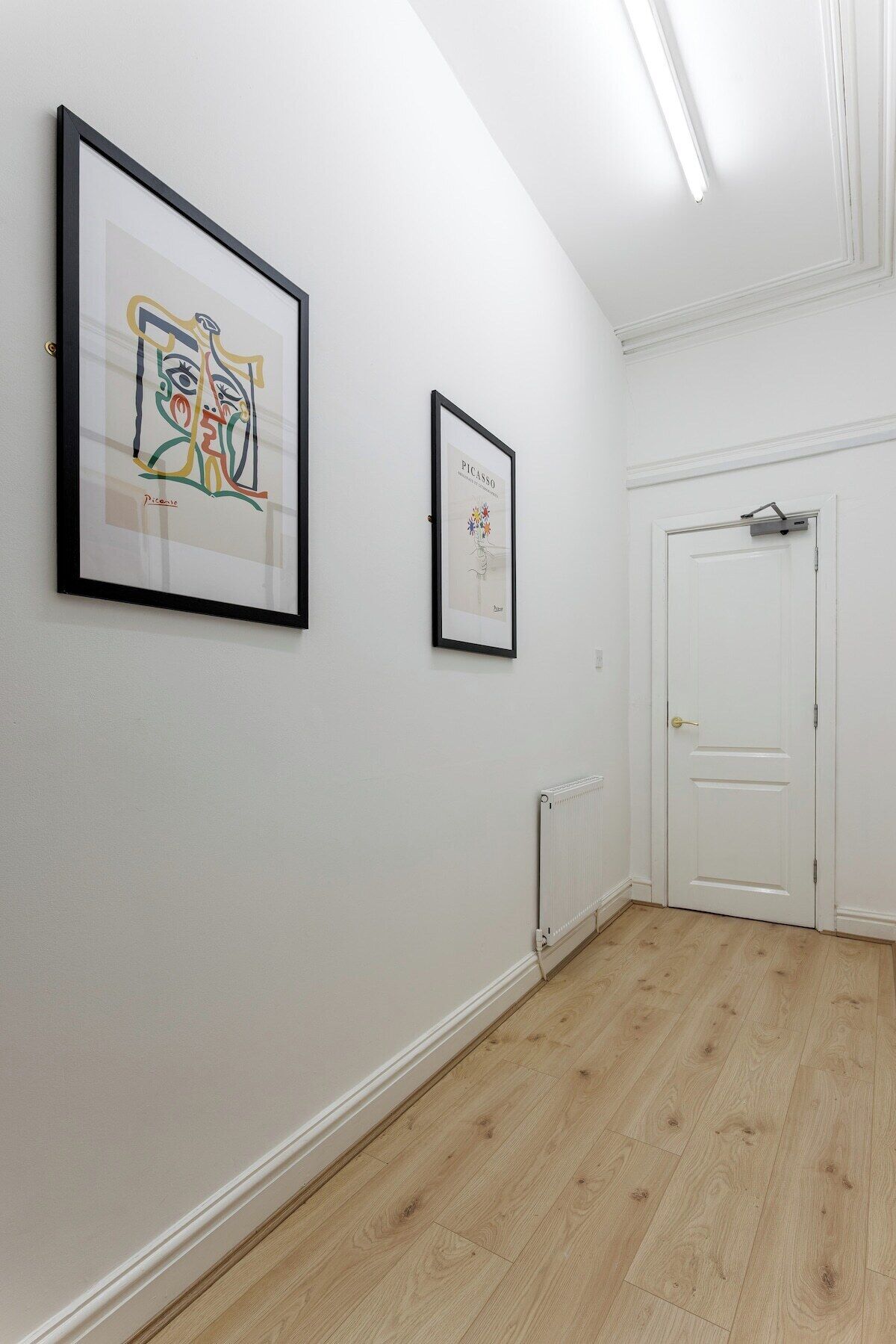 A hallway with two framed artworks on the wall, a radiator, and a white door.