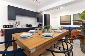 Une cuisine moderne avec des meubles de couleur sombre, une table à manger en bois pour quatre personnes et un coin salon avec un canapé et une grande plante.