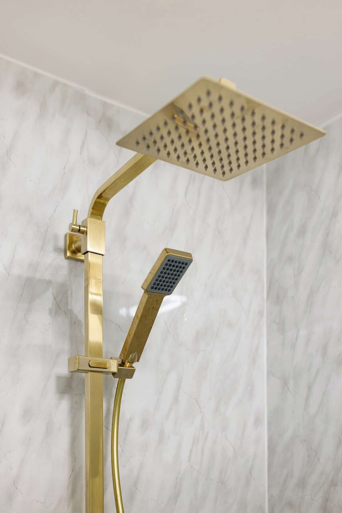 A gold-colored showerhead with a square design, mounted on a wall with a marble-like texture.