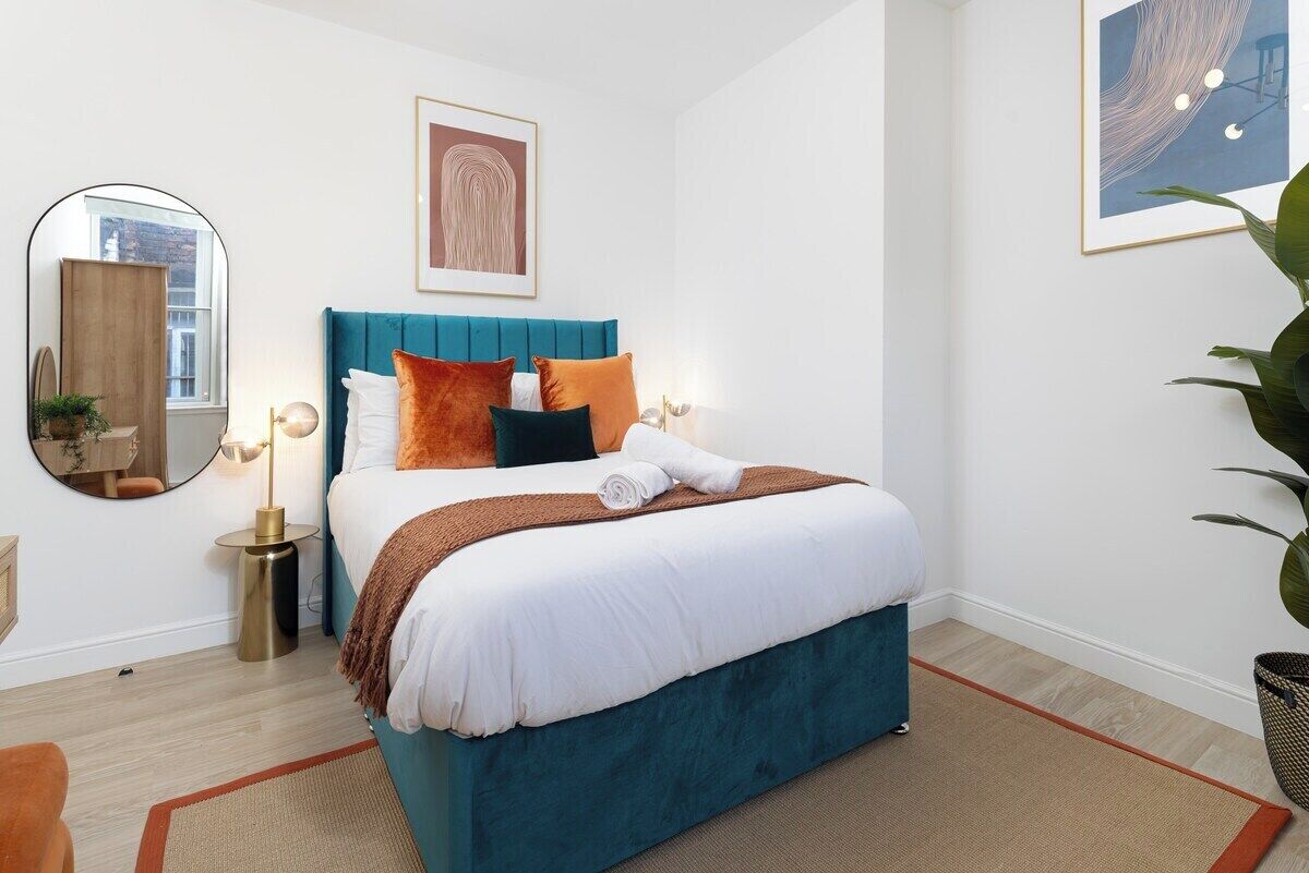 Un lit bien fait, avec une tête de lit bleue, un miroir rond et une œuvre d’art encadrée au-dessus.