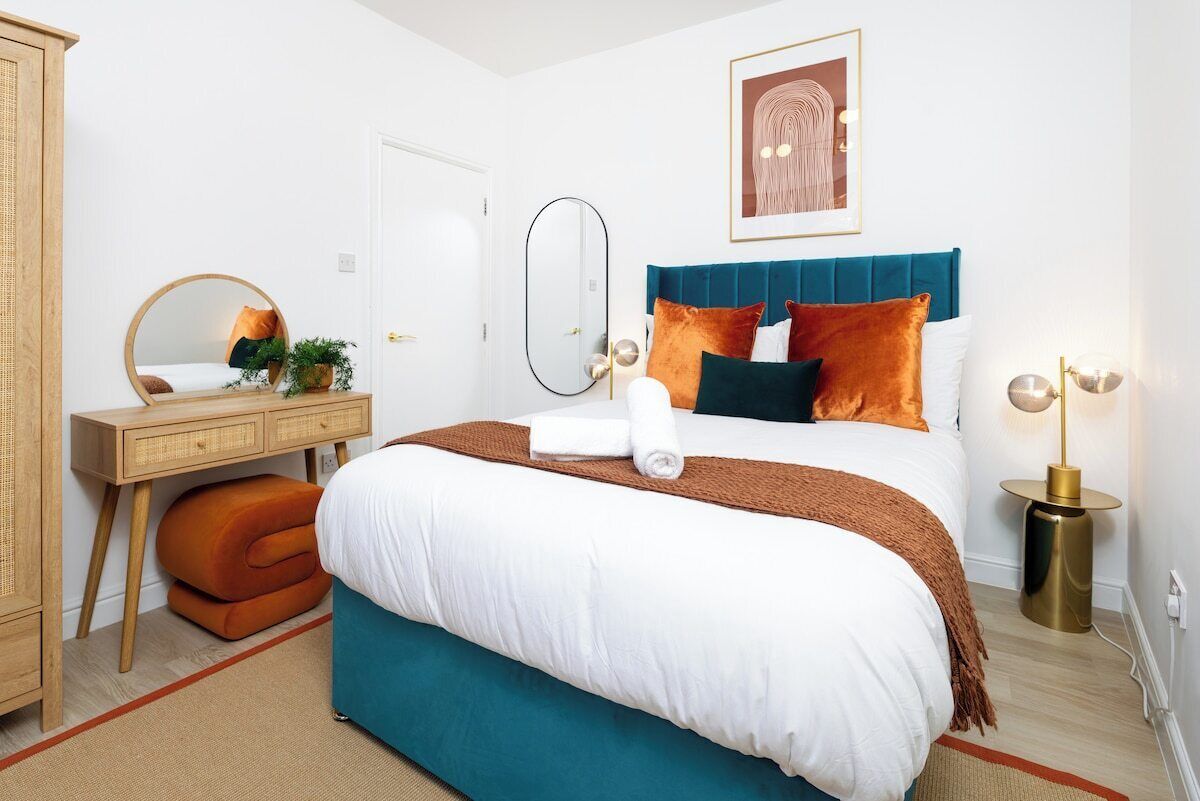 A hotel room with a bed, a desk, a mirror, a bedside table, and a wall-mounted artwork.