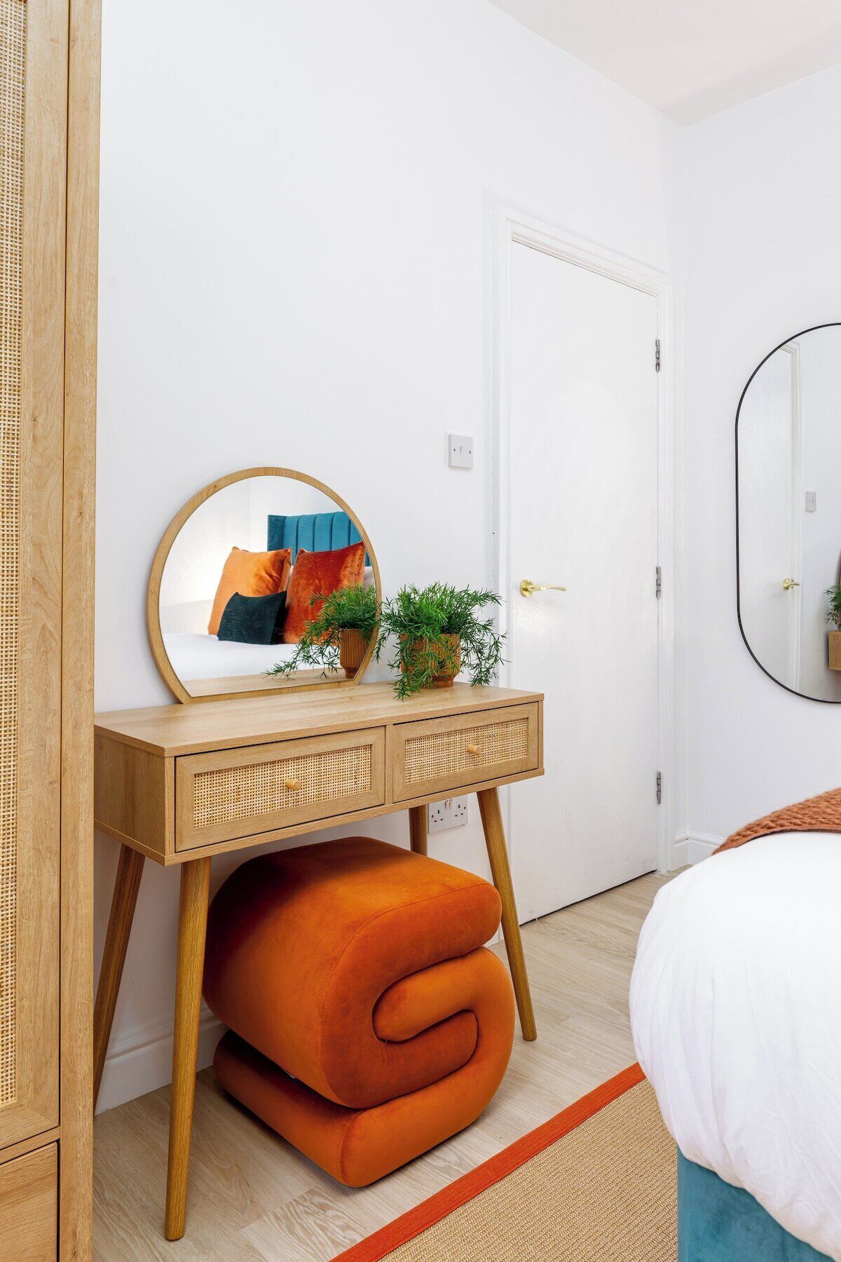 Une chambre avec une table de chevet en bois, un miroir, une chaise orange pliée et un lit avec une literie blanche.