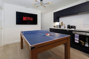 A room with a ping pong table, a TV displaying the Netflix logo, and a kitchen area with cabinets and appliances. - Flatzy City Centre Retreat  Sleeps 8  2 baths (Liverpool)