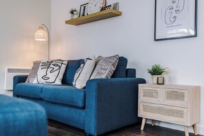 A modern living room with a blue sofa, patterned cushions, a floor lamp, a wooden side table, and a wall-mounted shelf with decorative items. - Flatzy - City Centre | 2 Bathrooms | Free Parking (Liverpool)