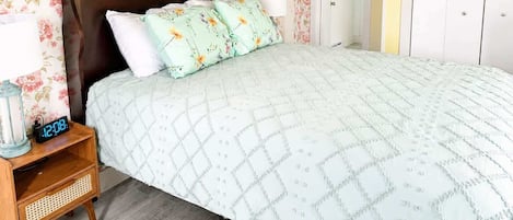 A bedroom with a floral wallpaper, a wooden headboard, a quilted bedspread, a bedside table with a lamp, and a clock.