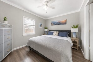 Une chambre à coucher comprenant un lit, un ventilateur de plafond, une fenêtre avec des stores, une table de chevet avec une lampe et une commode.