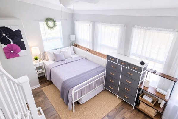 Une chambre à coucher avec un lit, une commode, une table de chevet, une lampe et une fenêtre décorée d’une guirlande.