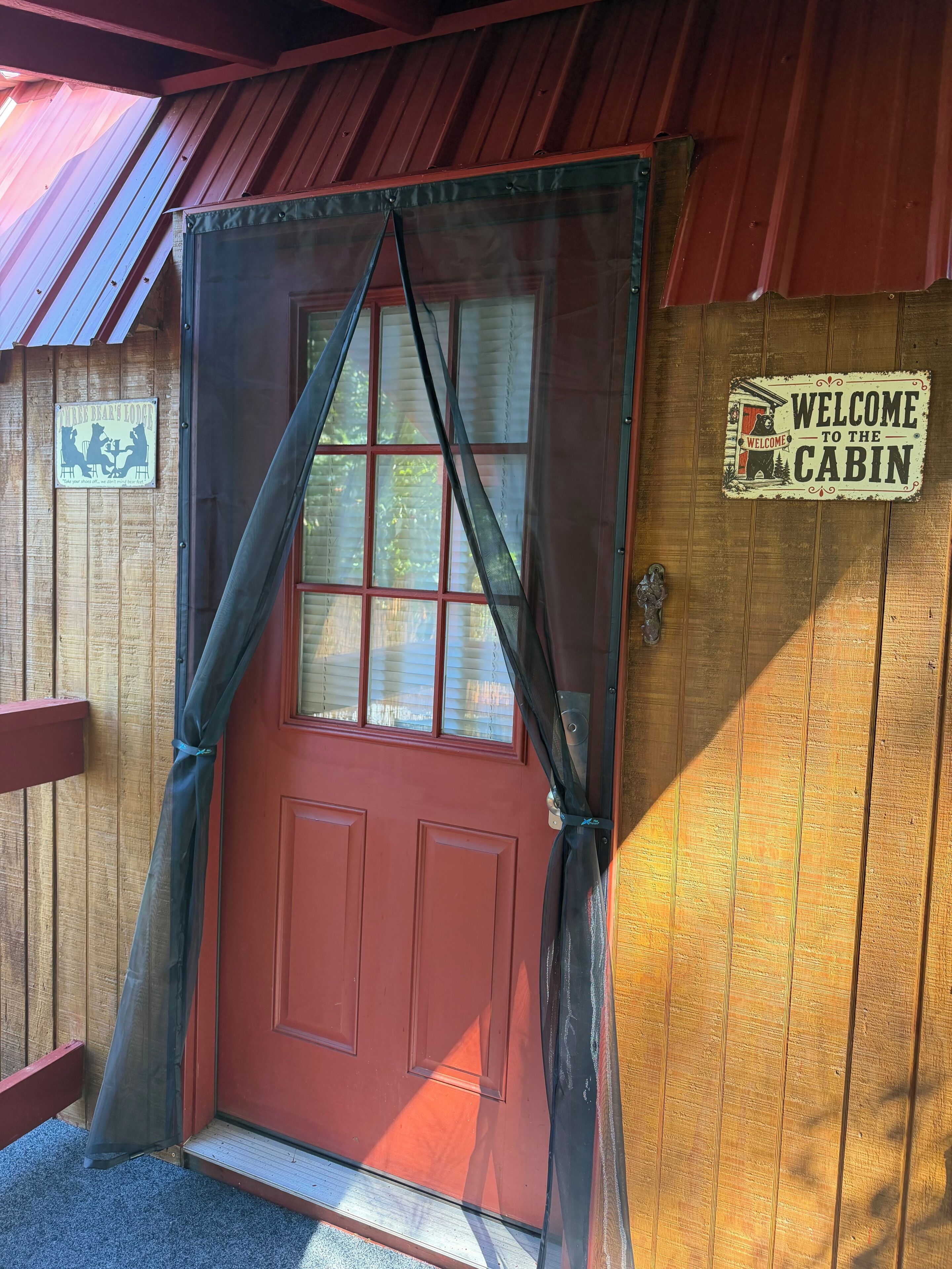 A red door with a mesh curtain, a welcome sign, and a wooden cabin exterior.