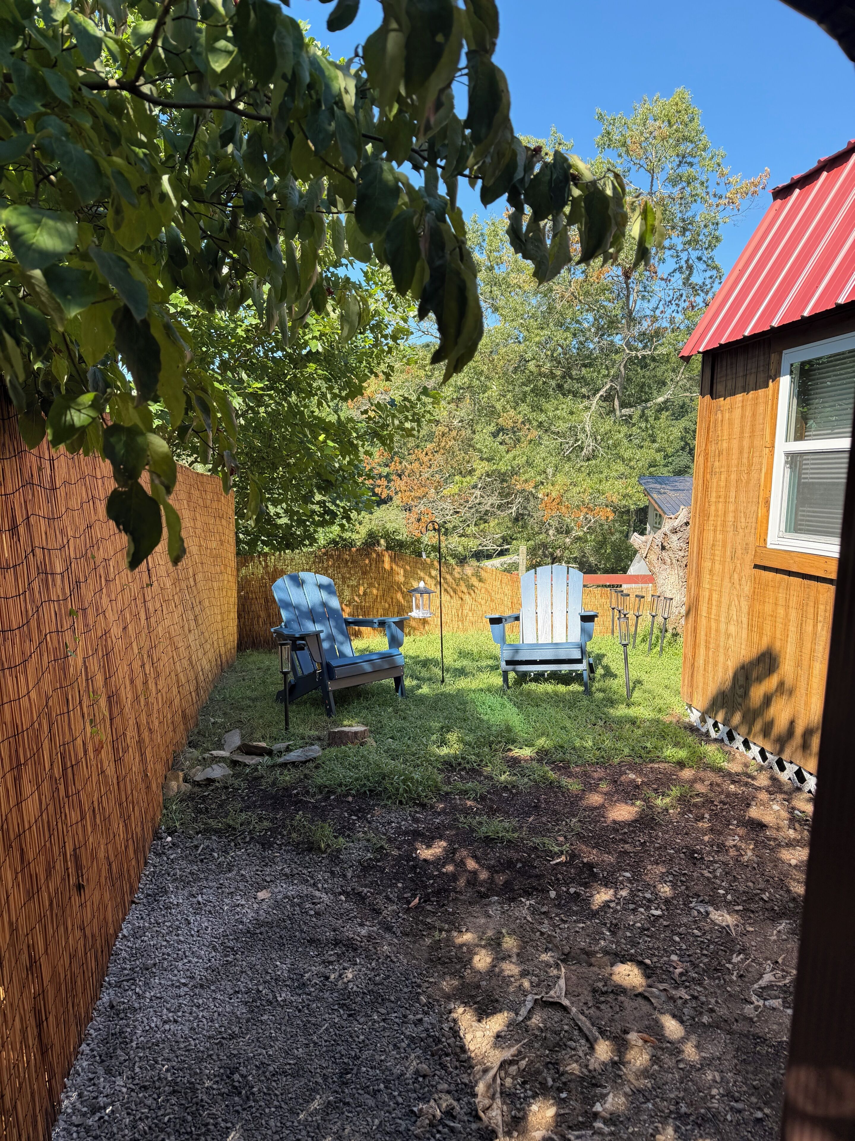 A backyard with a wooden fence, two Adirondack chairs, a small shed, and a red-roofed building.