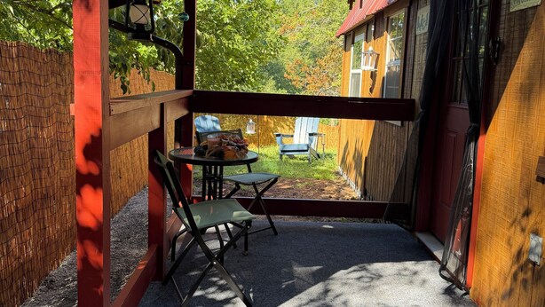 En overdækket terrasse med et bord og stole, et træhegn og udsigt til en hytte med rødt tag.