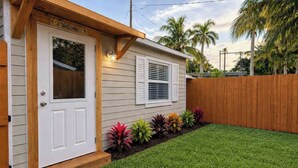 A single-story house with a white door and window shutters, a wooden porch, and a well-maintained lawn with colorful plants. - Lovely 1-Bedroom Studio in Jupiter Inlet Village (Jupiter)