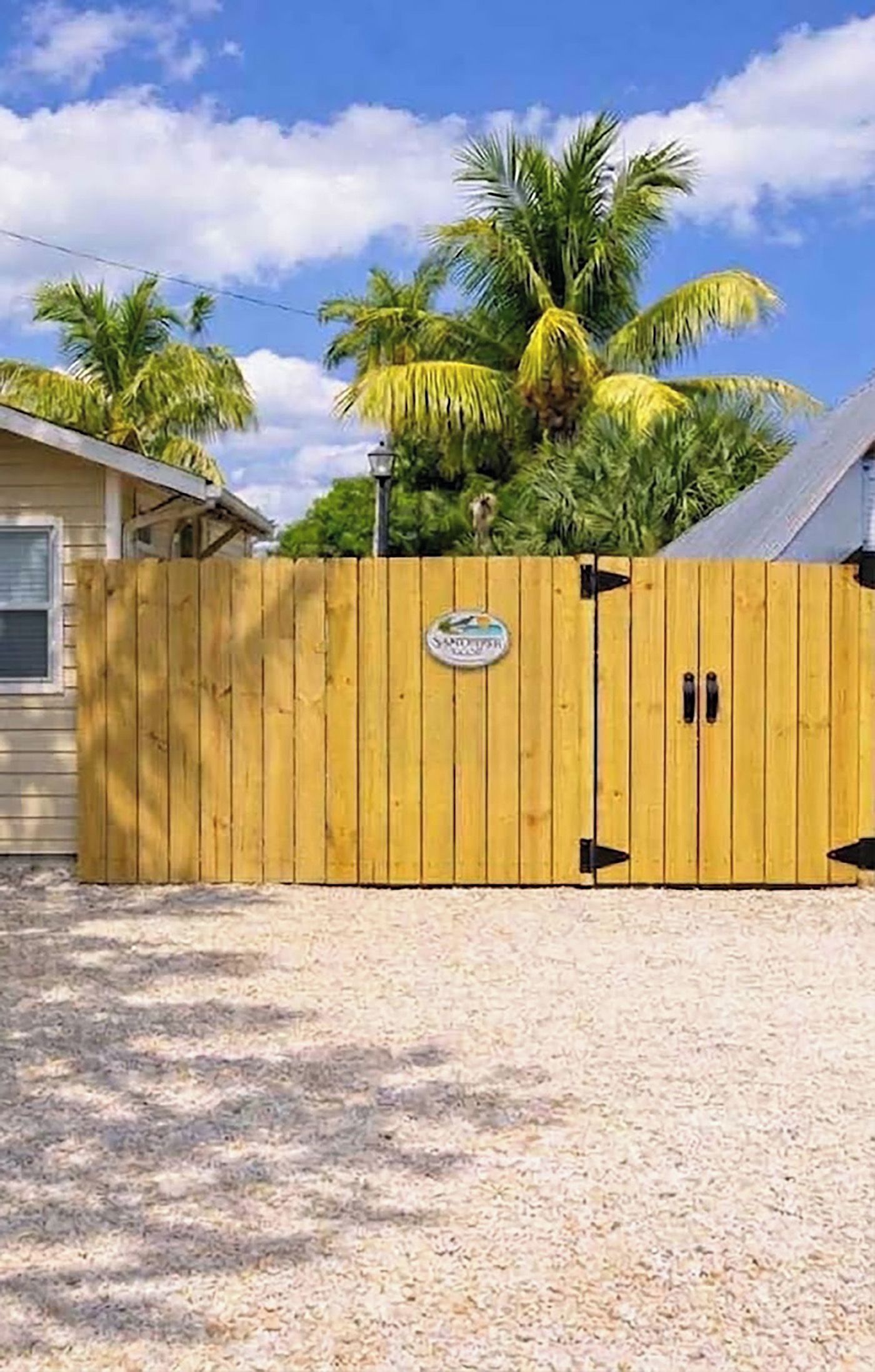 A wooden gate with a sign, a house, and palm trees in the background.