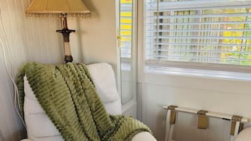 Ein Zimmer mit einem weißen Sessel, einer grünen Tagesdecke, einer Lampe und einem Fenster mit Jalousien.