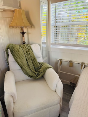 A room with a white armchair, a green throw blanket, a lamp, and a window with blinds.